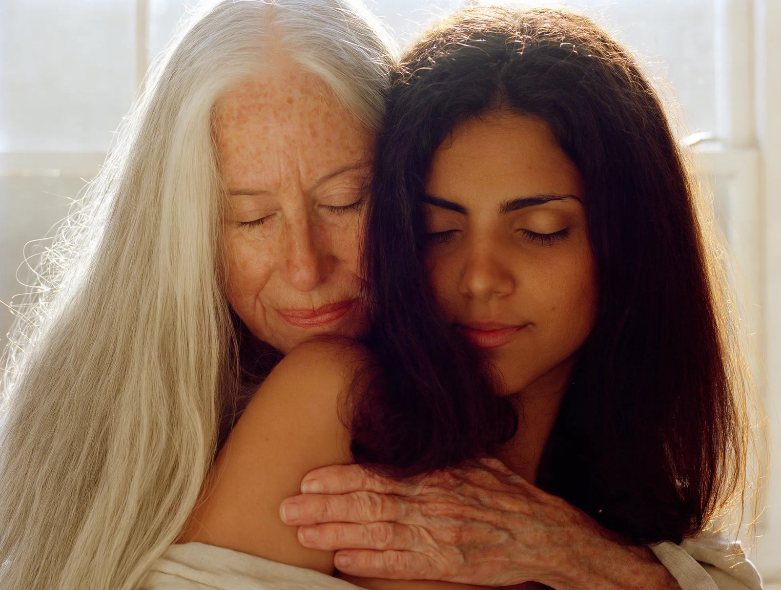 An older woman with long silver hair holding a younger woman with long dark hair, both with eyes closed
