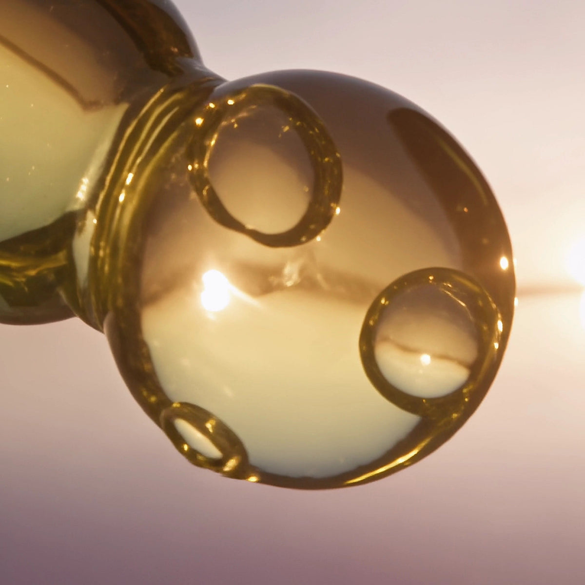 Macro of the glass pipette tip holding a round drop of the clear golden serum, tiny air bubbles inside, the low sun glowing through it