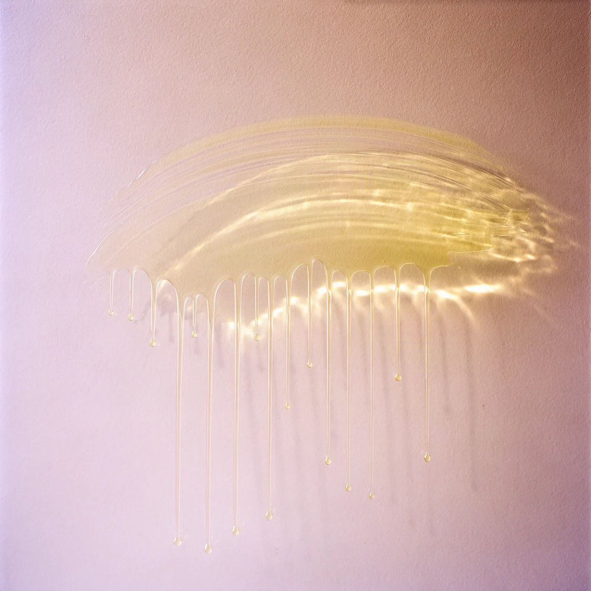 A thin sweep of the clear golden serum on a blush wall, fine drips running down, sunlight glowing through the oil