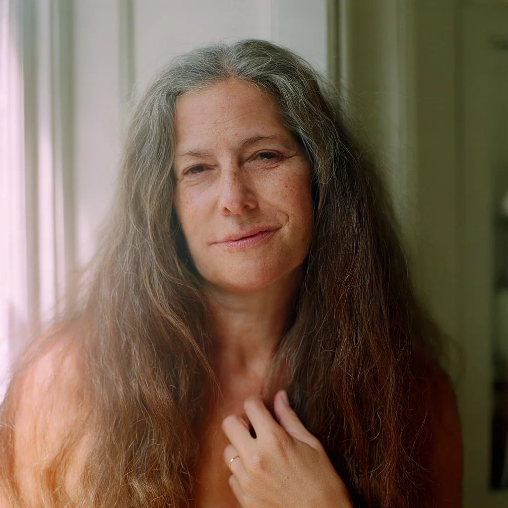 A woman with long silver-brown hair smiling, one hand resting at her chest