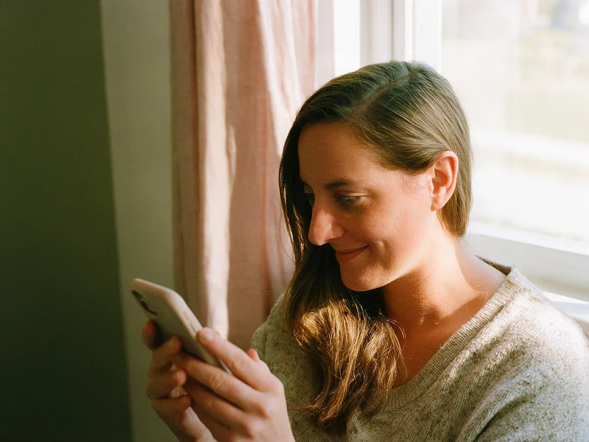 A woman by a window smiling softly at her phone